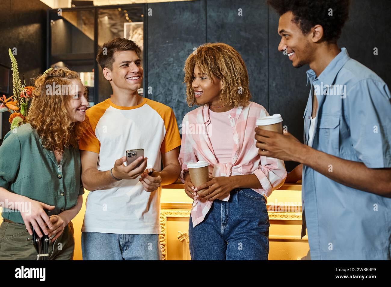 Junger Mann mit Smartphone lächelnd in der Nähe multiethnischer stilvoller Freunde an der Rezeption in einem modernen Hostel Stockfoto
