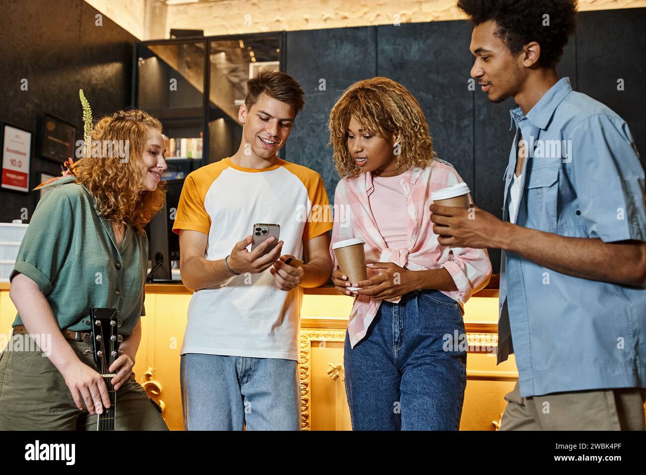 Glücklicher Mann, der multikulturellen Freunden Smartphone zeigt, mit Kaffee zum Mitnehmen und Gitarre in der Lobby des Hostels Stockfoto