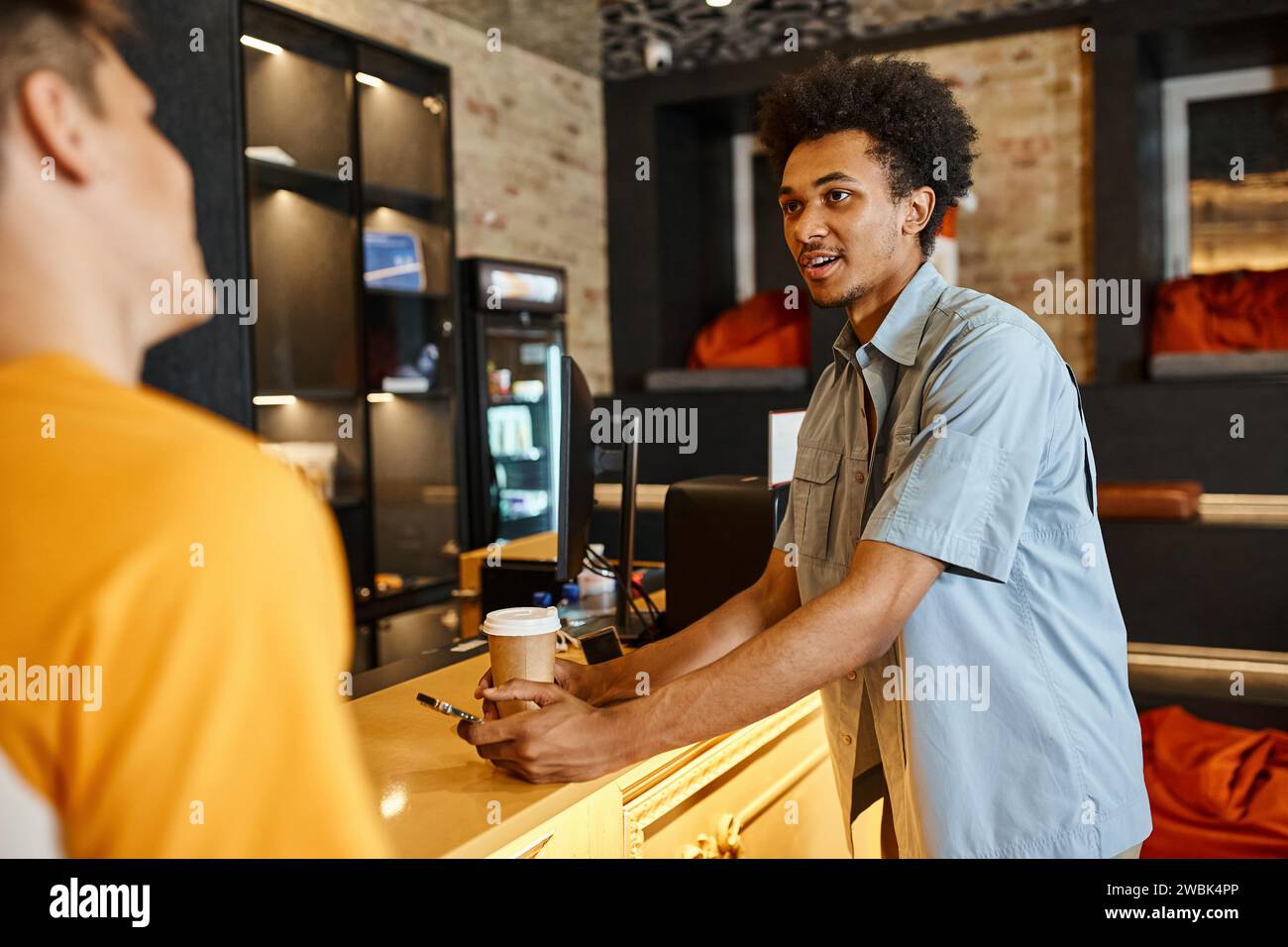 Junger afroamerikanischer Mann mit Smartphone und Pappbecher, der mit einem Freund in der Lobby der Jugendherberge spricht Stockfoto