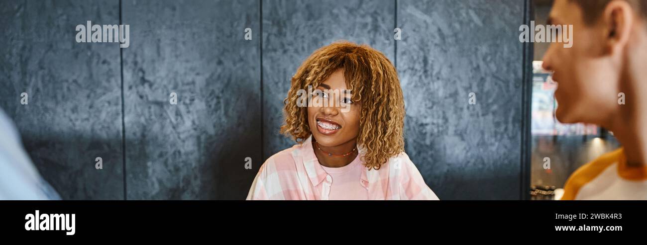 Jugendlicher und stilvoller afroamerikanischer Rezeptionist lächelt in der Nähe von Studenten in der Jugendherberge, Banner Stockfoto