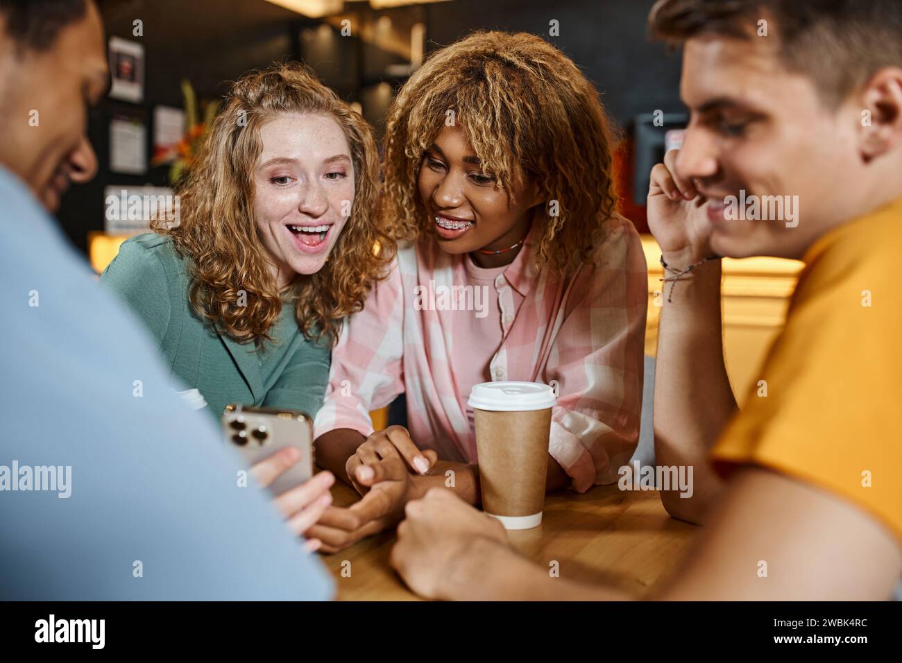 Aufgeregte Frau, die glücklichen multiethnischen Freunden im Lounge-Café der Studentenherberge ihr Smartphone zeigt Stockfoto