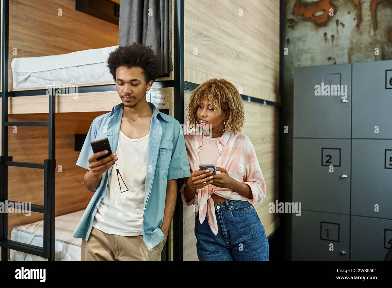 Trendige afroamerikanische Studenten, die Smartphones in der Nähe von Doppeldeckerbetten in einem modernen Hostel nutzen Stockfoto