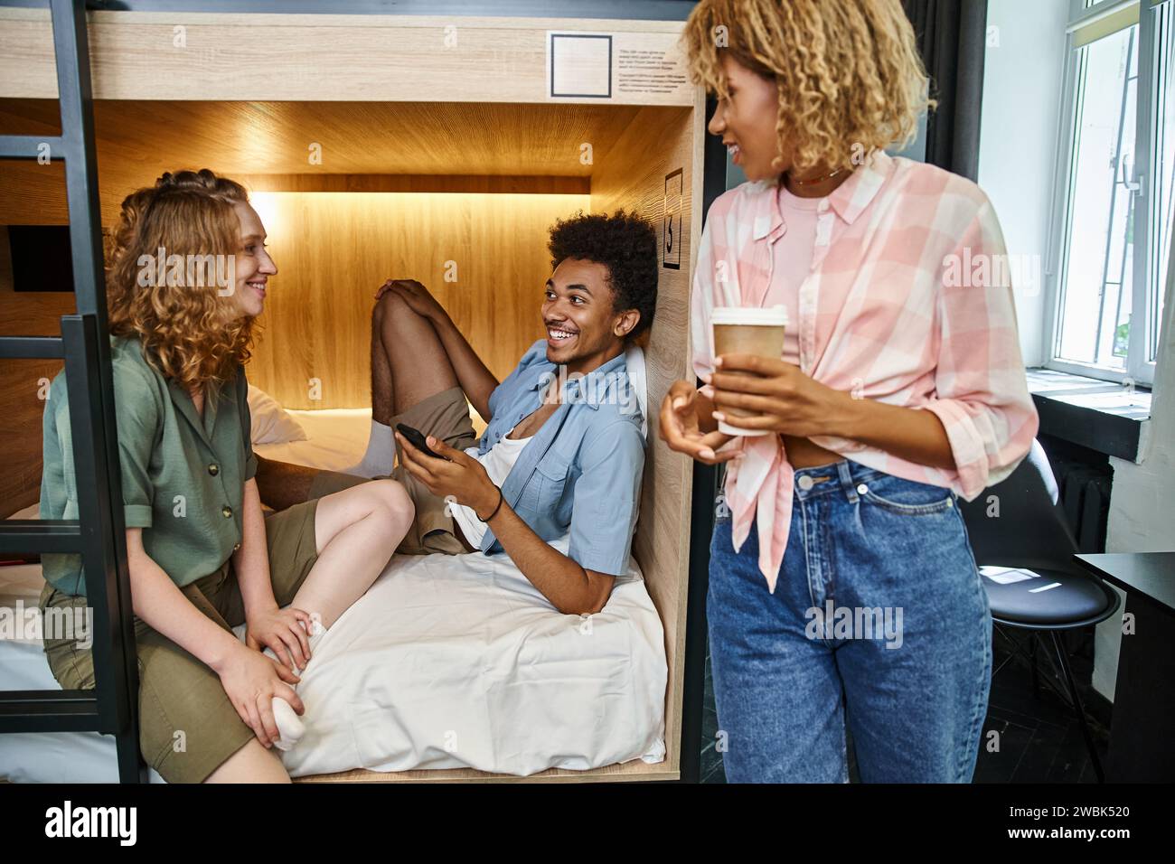 Fröhliche afrikanische Studentin mit Smartphone auf Doppeldeckerbett in der Nähe weiblicher Freunde in Jugendherberge Stockfoto