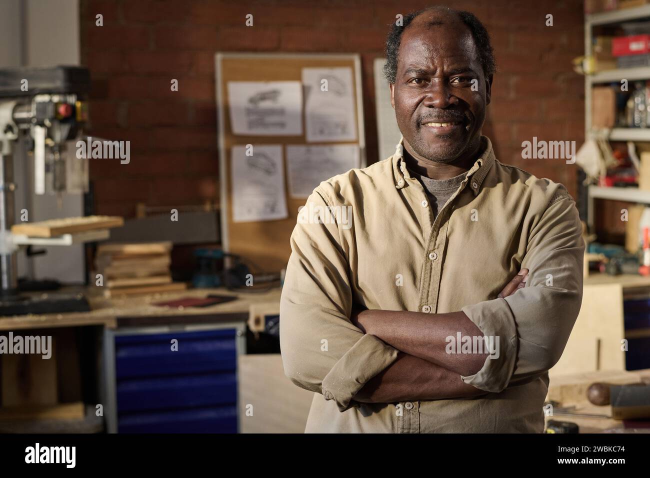 Porträt eines älteren schwarzen Zimmermanns, der die Kamera anlächelt und die Arme in der Werkstatt überkreuzt Stockfoto