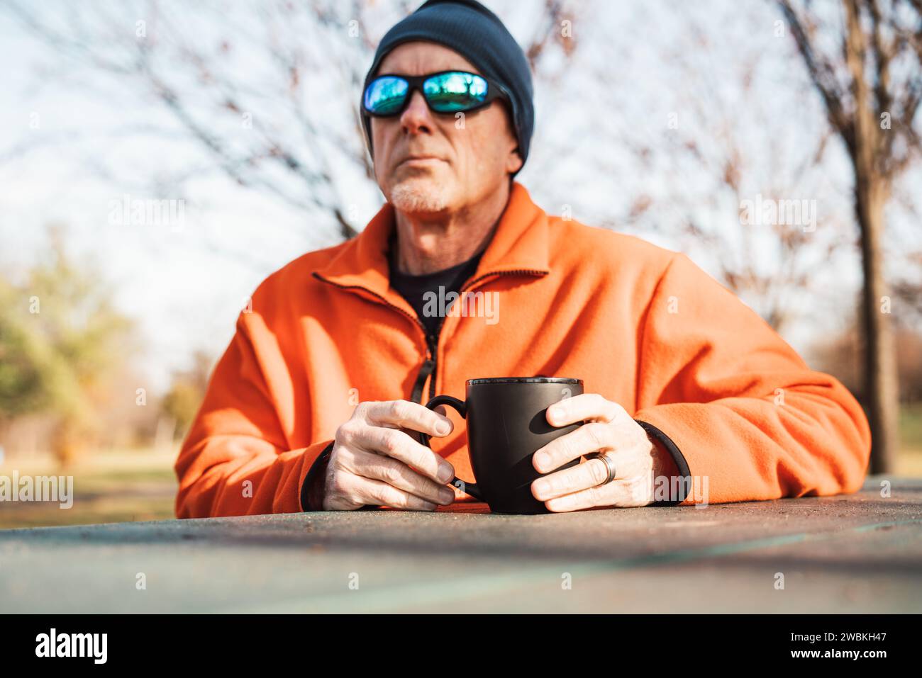 Ein älterer Mann mit Sonnenbrille sitzt an einem Tisch und genießt bei kaltem Wetter draußen eine Tasse Kaffee Stockfoto