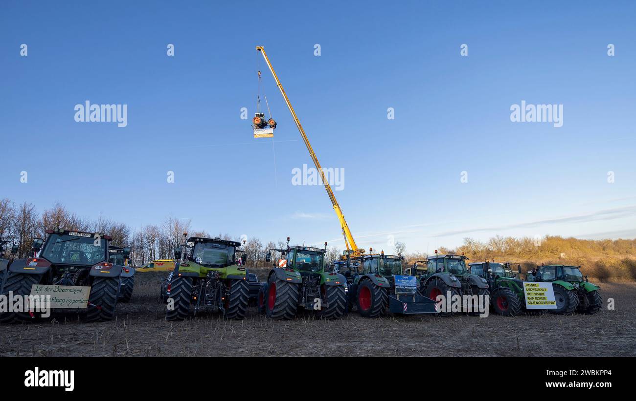 Mainstockheim, Deutschland. Januar 2024. Ein Traktor mit der Aufschrift „Landwirtschaft am Faden“ wird am Autobahnkreuz Biebelried auf der Deponie LZR DK0 in Mainstockheim, wo andere Traktoren abgestellt sind, aus Protest gegen die Sparpläne der Bundesregierung von einem Kran hochgezogen. Ein Banner lautet: "Wenn es keine Landwirtschaft gibt, kann es auch keinen Staat geben". Quelle: Heiko Becker/dpa/Alamy Live News Stockfoto