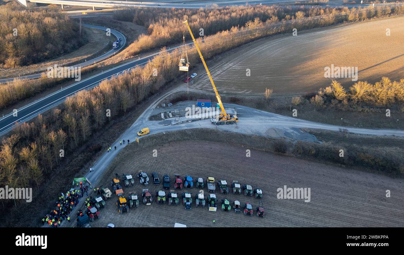 Mainstockheim, Deutschland. Januar 2024. Ein Traktor mit der Aufschrift „Landwirtschaft am seidenen Faden“ wird am Autobahnkreuz Biebelried an der Deponie LZR DK0 in Mainstockheim, wo andere Traktoren abgestellt sind, aus Protest gegen die Sparpläne der Bundesregierung von einem Kran hochgezogen. (Foto mit einer Drohne) Credit: Heiko Becker/dpa/Alamy Live News Stockfoto