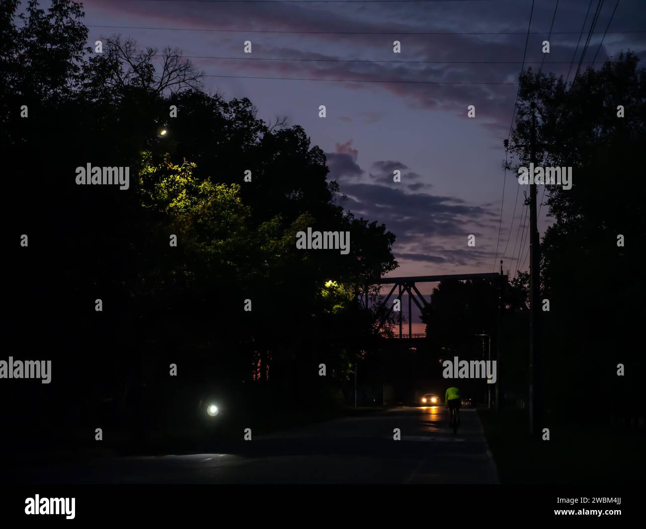 Radfahrer fahren mit dem Fahrrad auf einem Fahrradweg in Richtung einer Brücke und einem wunderschönen farbenfrohen Sonnenuntergang. Auto mit Scheinwerfer an, Bäume mit Straßenlaternen beleuchtet Stockfoto