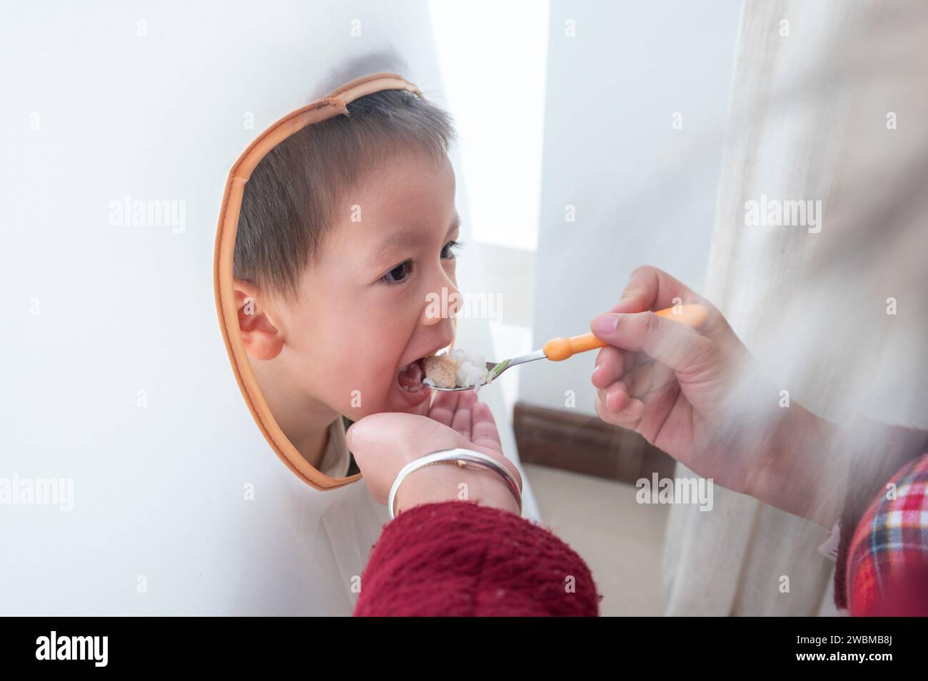 Der süße kleine Junge blickt aus dem Fenster eines weißen Kinderzelt, während seine Mutter ihn mit Löffel füttert, um ihn zum Mittagessen am Tisch einzuladen Stockfoto