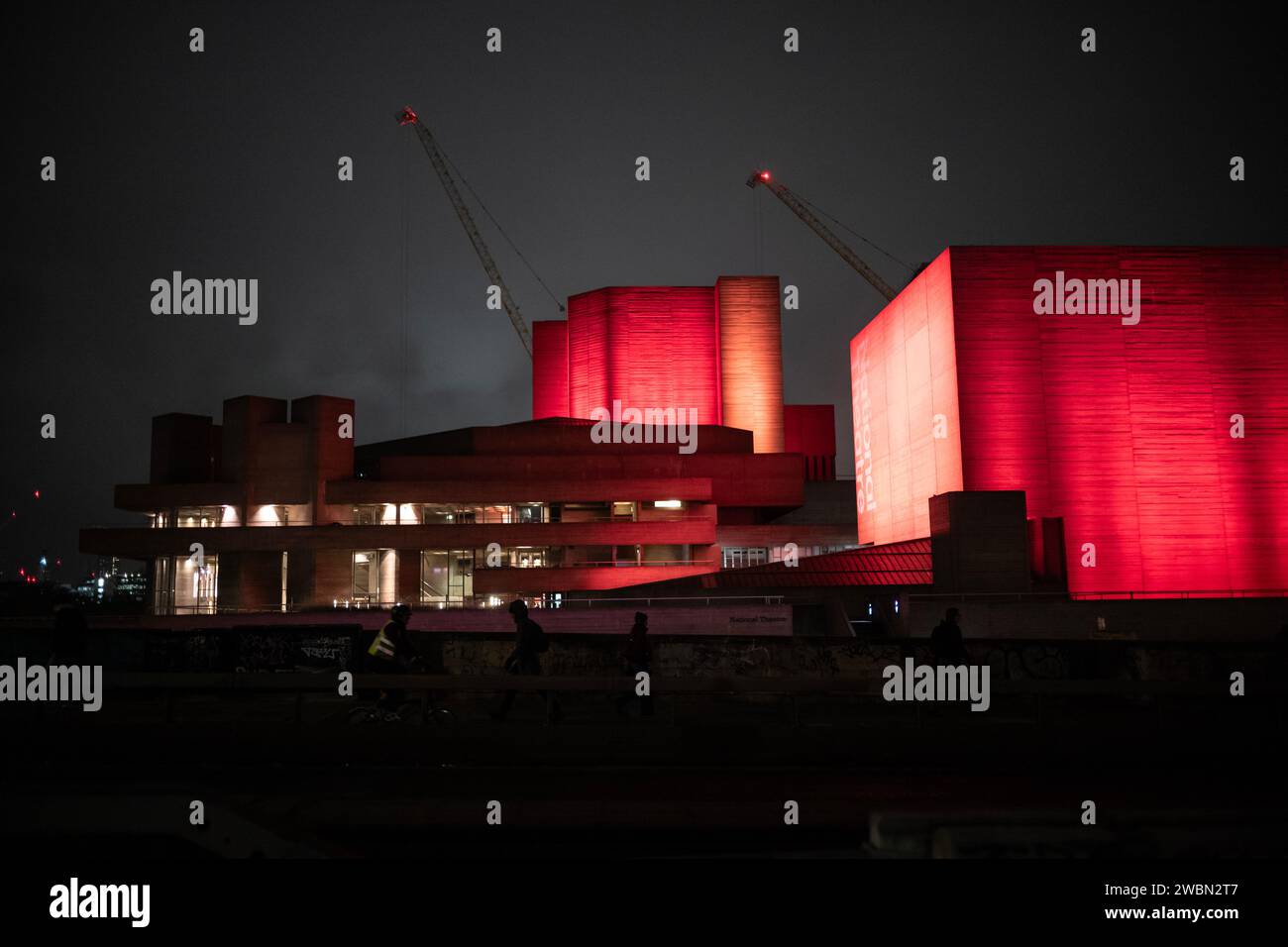 Pendler laufen in einer Winternacht über die Waterloo Bridge neben einem roten Ufer des National Theatre, Southbank, im Zentrum von London, England. Stockfoto