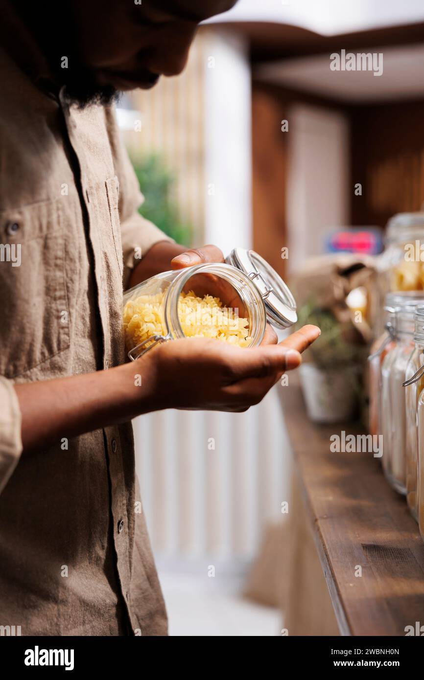 Nahaufnahme eines schwarzen Mannes, der frisches, lokal angebautes Getreide und Nudeln untersucht. Selektiver Fokus eines afroamerikanischen männlichen Kunden, der ein Glas mit Spaghetti hält und das hausgemachte Produkt bewundert. Stockfoto