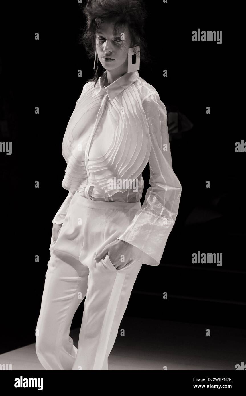Modemodell mit transparenter weißer Langarmbluse mit weißen Nadelstreifen und einer großen runden Stoffapplikation und weißer Hose auf der Runway Fashion Show Stockfoto