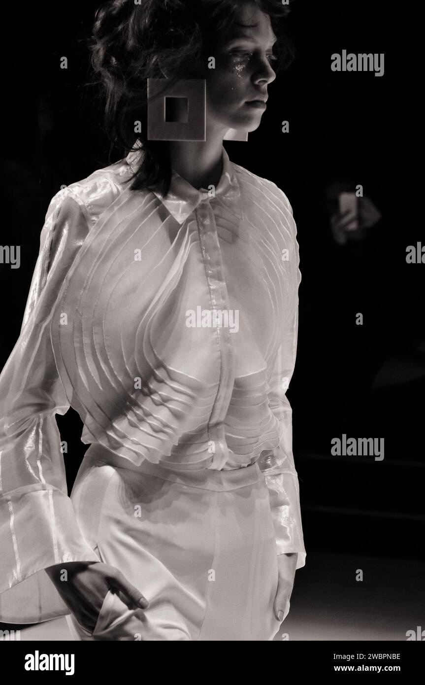 Model mit transparenter weißer Langarmbluse mit weißen Nadelstreifen, großer, runder Stoffapplikation und weißer Hose auf der Runway Fashion Show Stockfoto