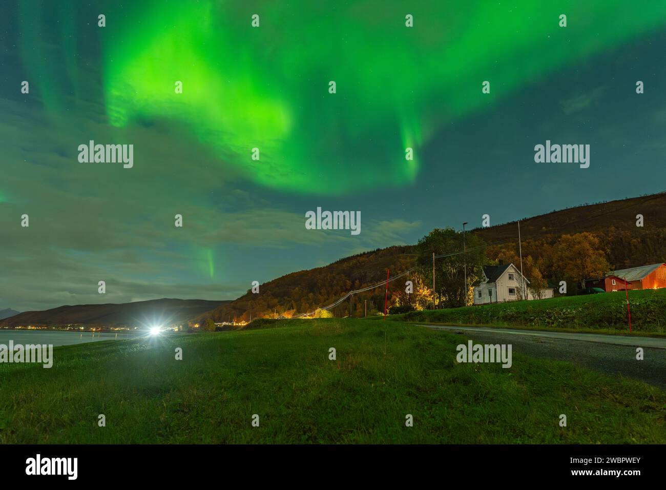 Wunderschöne grüne Nordlichter über einem Fjord und einem Haus auf der Insel Kvaloya in der Nähe von Tromsø. Tanzende Polarlichter über einem Berg, aurora borealis Stockfoto