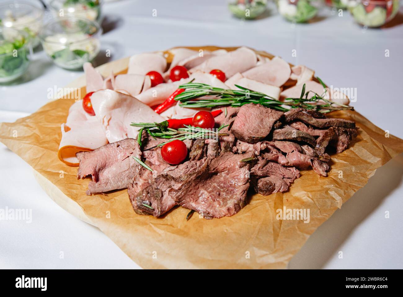 Eine Gourmet-Fleischplatte mit saftig geschnittenem Roastbeef und truthahn, garniert mit Kirschtomaten und frischem Rosmarin auf einem rustikalen, papierbeschichteten Holz Stockfoto