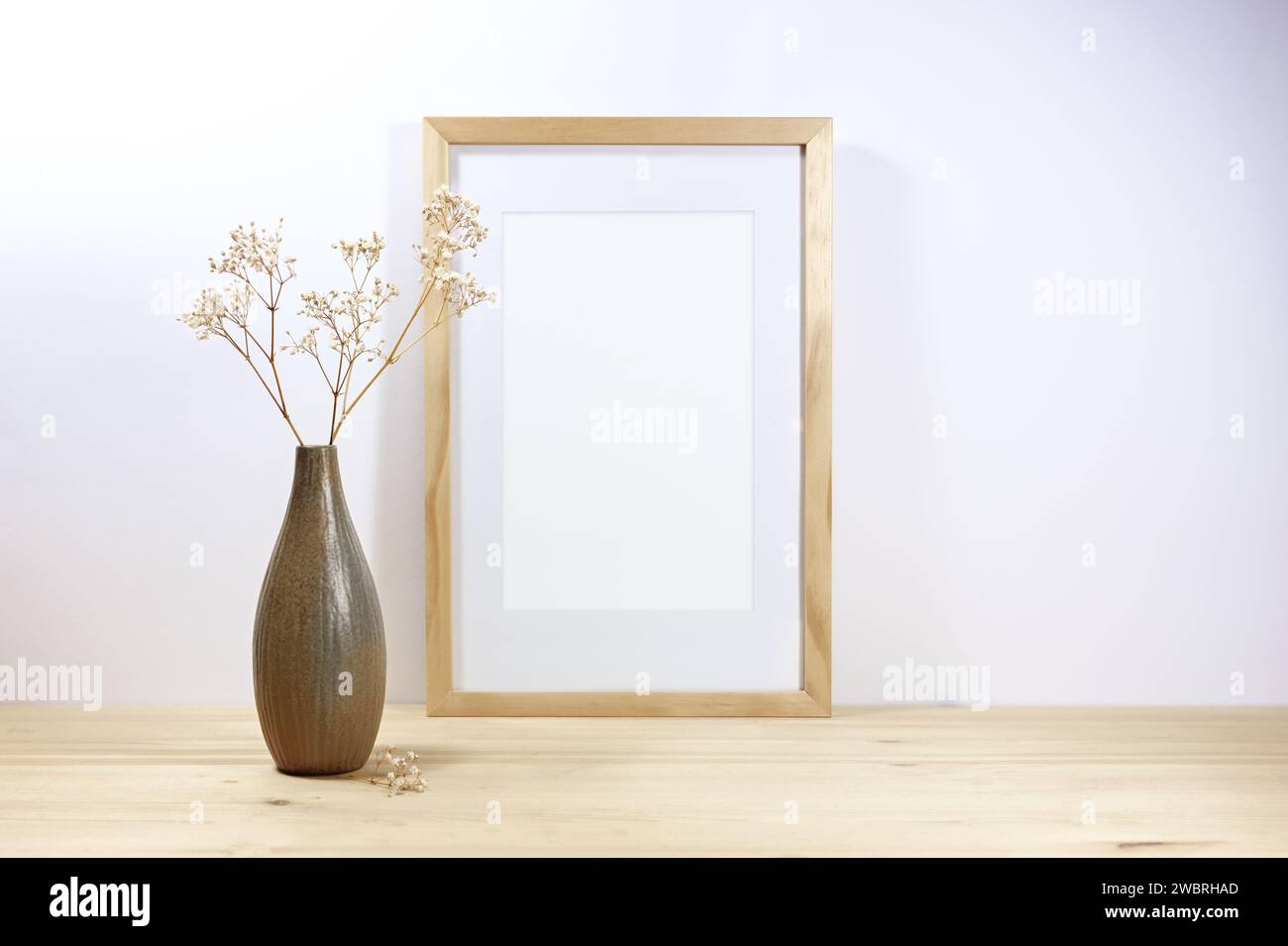 Leeres Bilderrahmenmockup und eine braune Keramikvase mit filigraner gypsophila auf einem Holztisch vor einer hellgrauen Wand, minimales Wohndekor, Skandina Stockfoto
