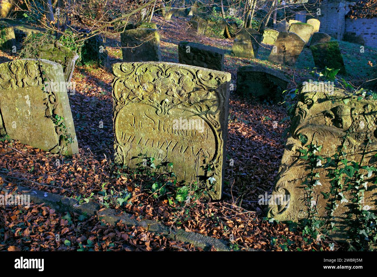 Die Wintersonne am späten Nachmittag beleuchtet kunstvolle Details auf alten verwitterten Grabsteinen auf einem Kirchengatter. Stockfoto