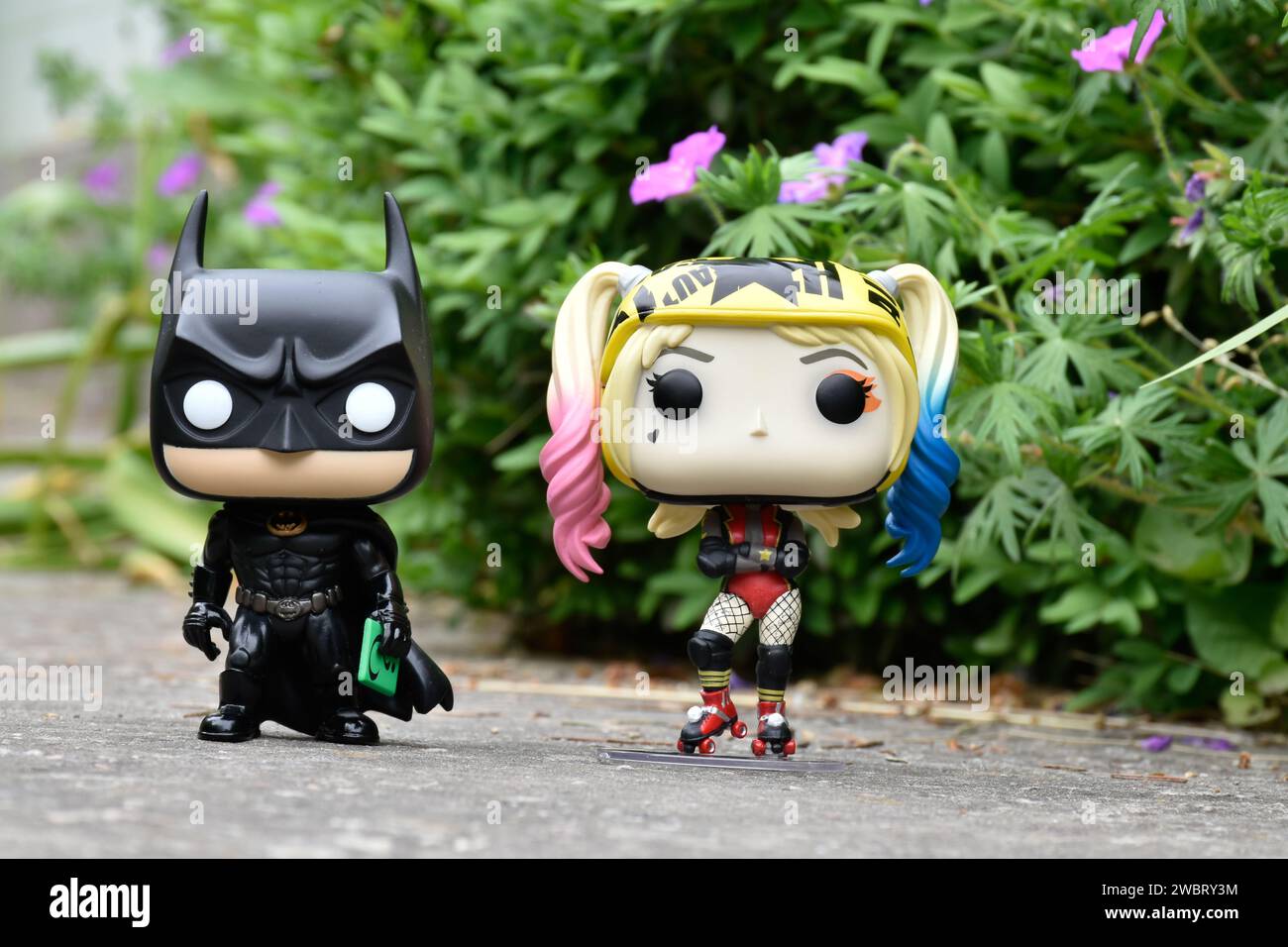 Funko Pop Actionfiguren der DC-Comic-Superhelden Batman und Harley Quinn stehen auf Asphaltstraße im Frühlingspark, grünes Gras und rosa Blumen. Stockfoto