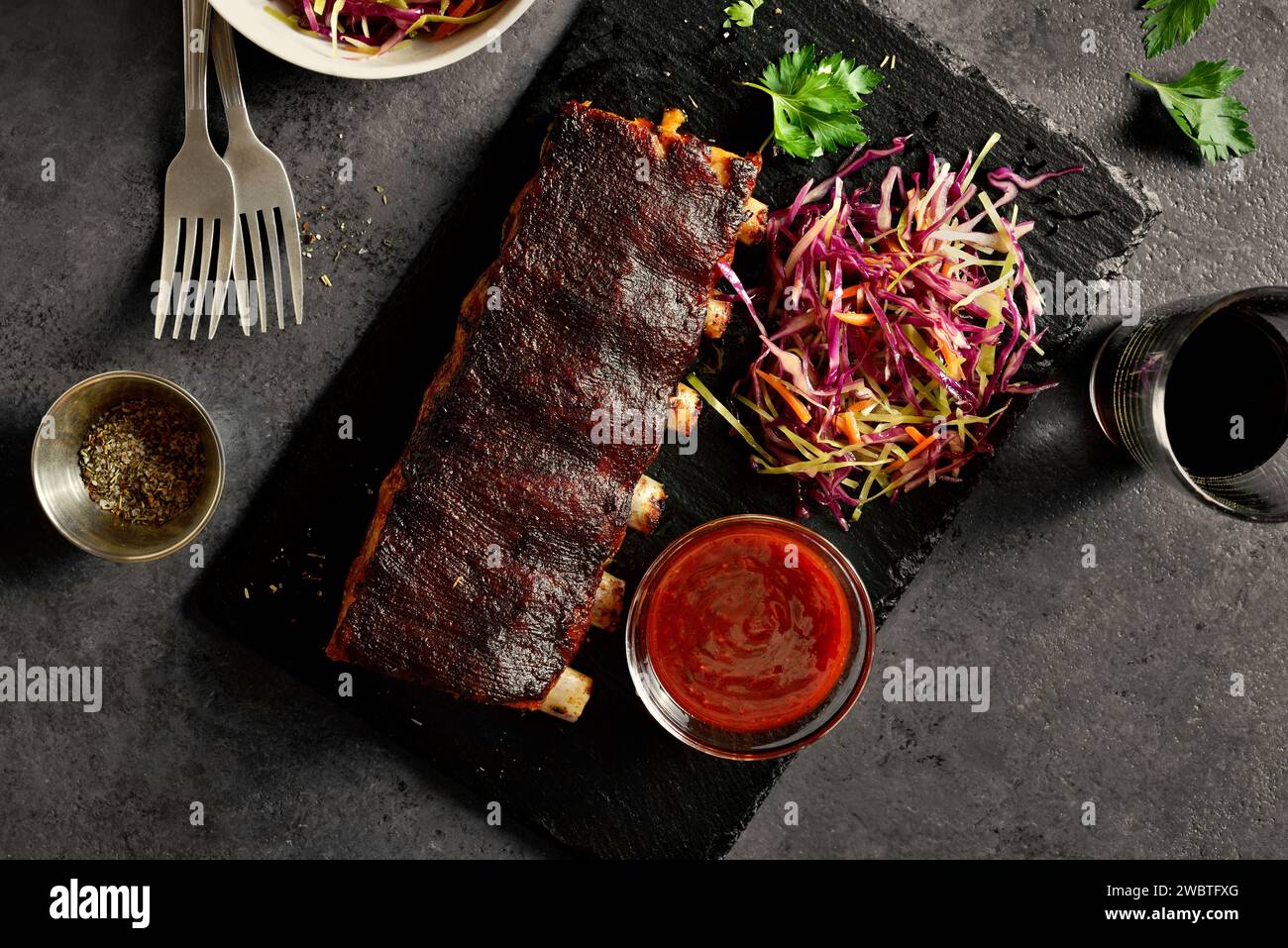 Gegrillte Schweinerippchen, serviert mit Krautsalat und Sauce auf schwarzem Schieferteller auf dunklem Hintergrund. Nahaufnahme von bbq Schweinerippchen, serviert mit lebhaften coles Stockfoto