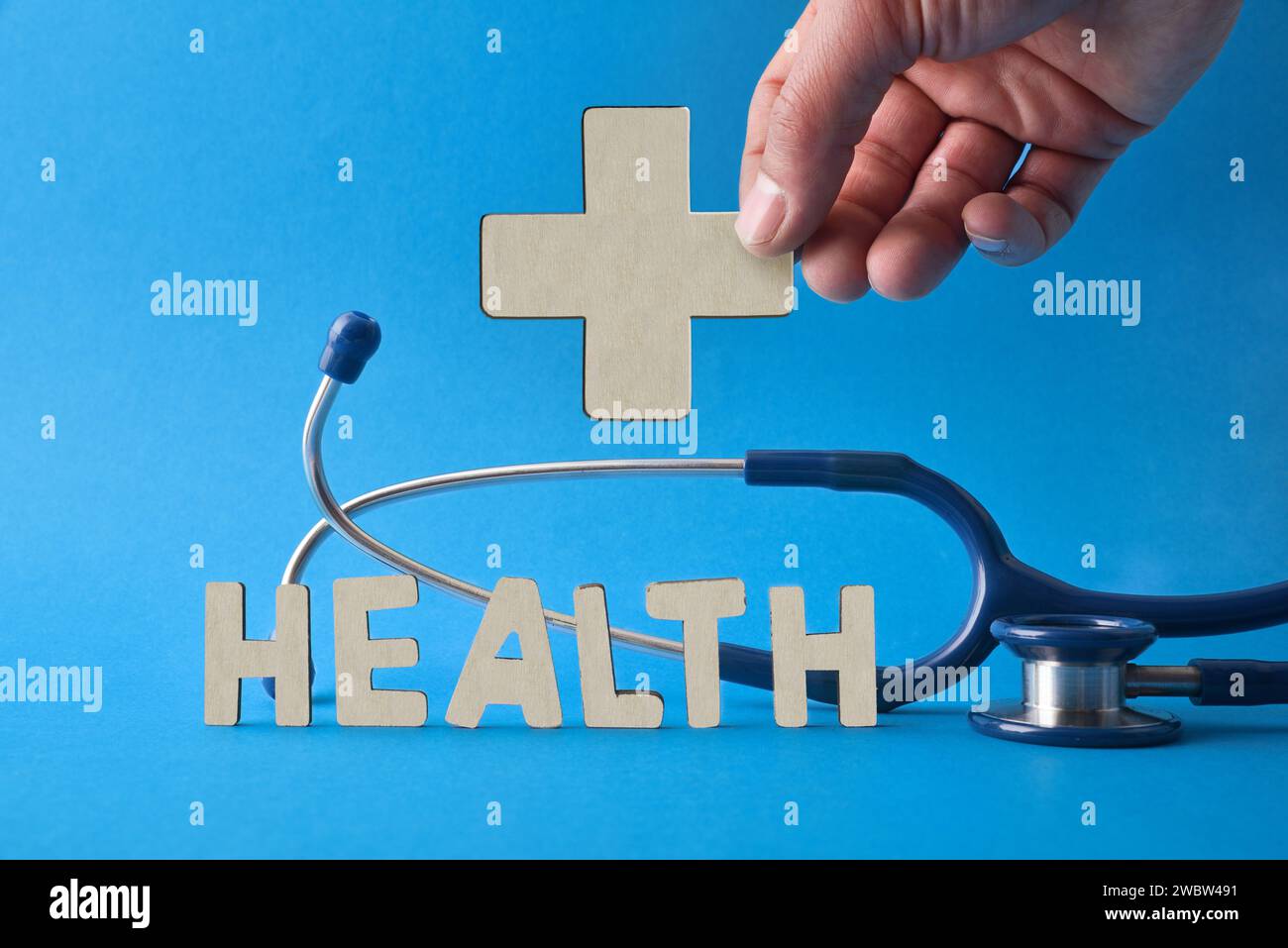 Medizinischer Gesundheitshintergrund mit Hand haltendem Kreuz und hölzernen Buchstabenausschnitten auf blauem Tisch und Stethoskop mit blauem Hintergrund. Vorderansicht Stockfoto