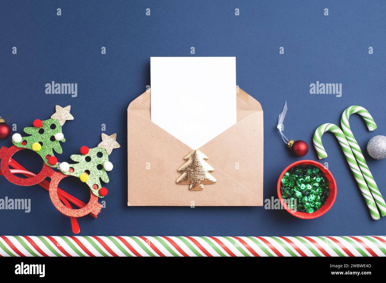 Draufsicht des Umschlags mit leerem Papier, Geschenkpapier, grünem Glitzer, Pailletten, Süßigkeiten, Weihnachtskugeln, Dekorationen auf blauem Hintergrund. Stockfoto