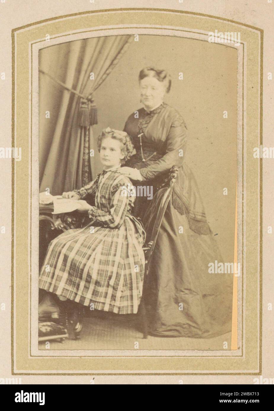 Porträt eines Mädchens in einem Kleid mit schottischem Fenster und einer Frau in einem Kleid an einem Tisch mit Büchern, Anonym, ca. 1860 - ca. 1900 Fotografie. Besuchskarte Teil des Fotoalbums einer niederländischen Familie mit 168 Cartes de-Visiting- und Kabinettfotos. Pappe. Papier. Fotografischer Trägeralbumendruck historischer Personen. Kleid, Kleid. studiobedarf  Fotograf. Friseurstile - AA -  Frauen. Kind Stockfoto
