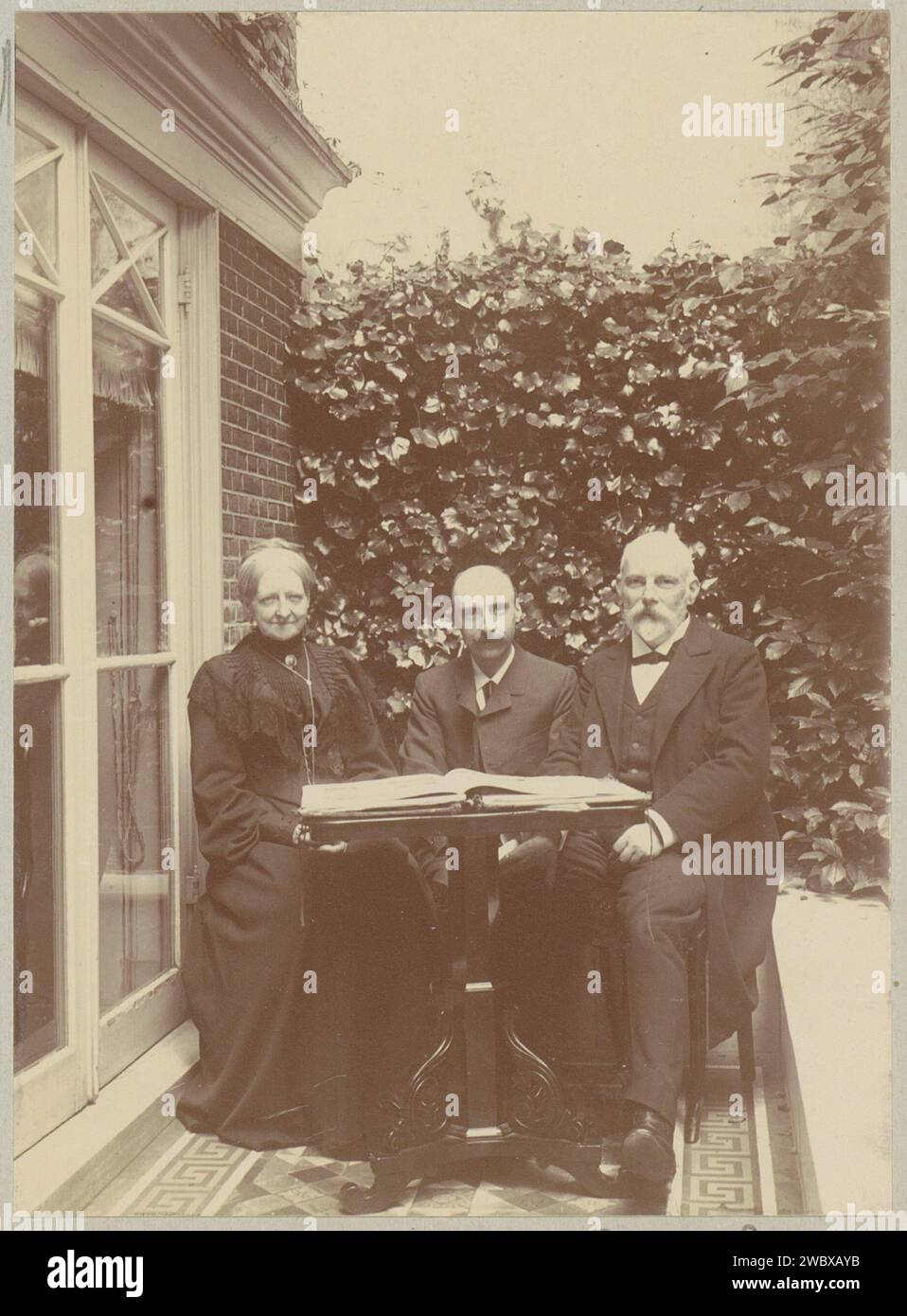 Zwei Männer und eine Frau sitzen in einem Haus mit einem Buch auf dem Tisch, anonym, ca. 1900 - ca. 1910 Foto Teil des Familienalbums mit Fotos von Wijnhandel Kraaij & Co. Bordeaux-Amsterdam. Fotografische Unterstützung beim Lesen Stockfoto