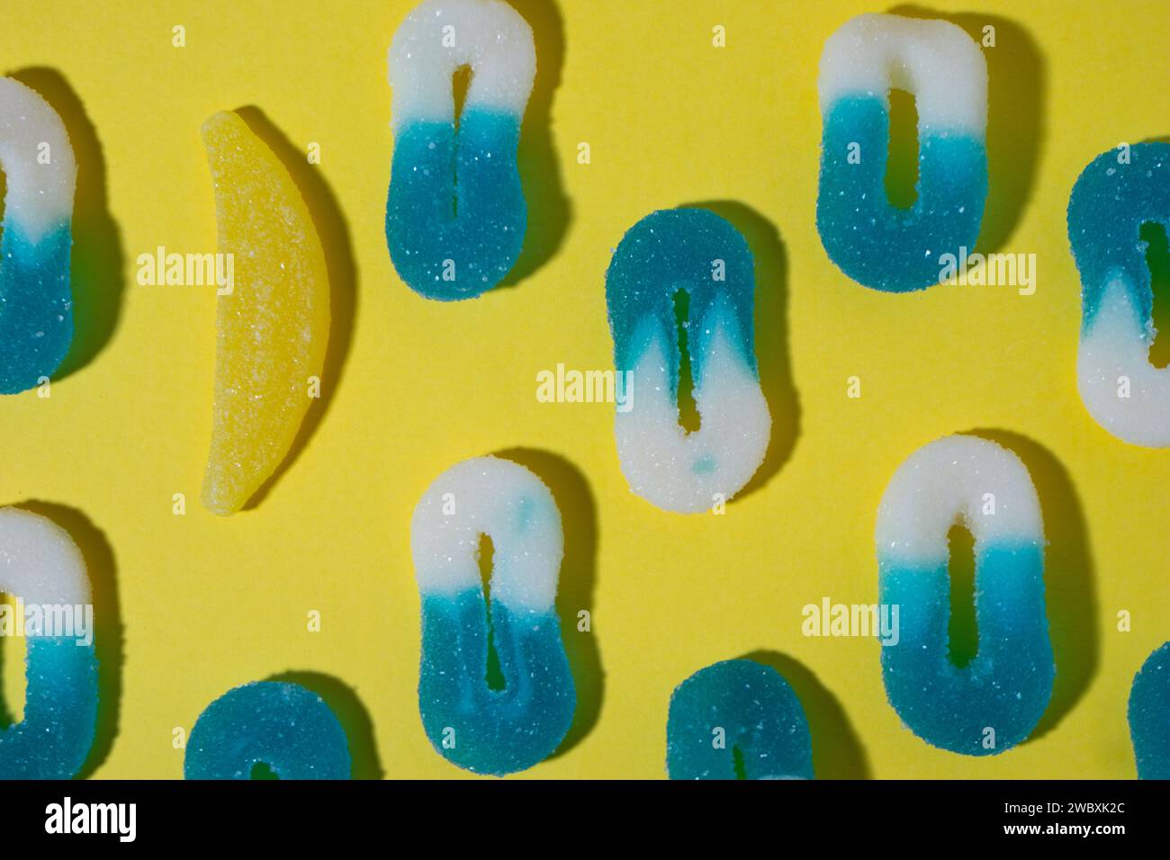 Anordnung von 0-förmigen weißen und blauen gummiartigen Bonbons auf gelbem Hintergrund, von denen eine durch einen Bananengummi ersetzt wird, in einem horizontalen Bild W Stockfoto