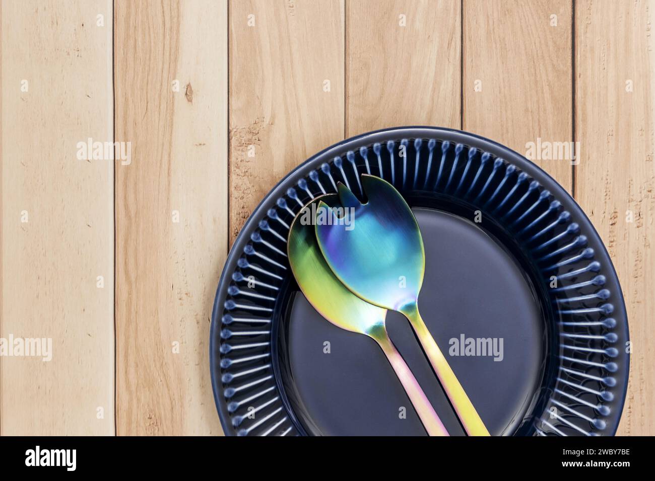 Ein hellblauer Porzellanteller und Salatbesteck aus Metall auf einem hellen Holztisch Stockfoto