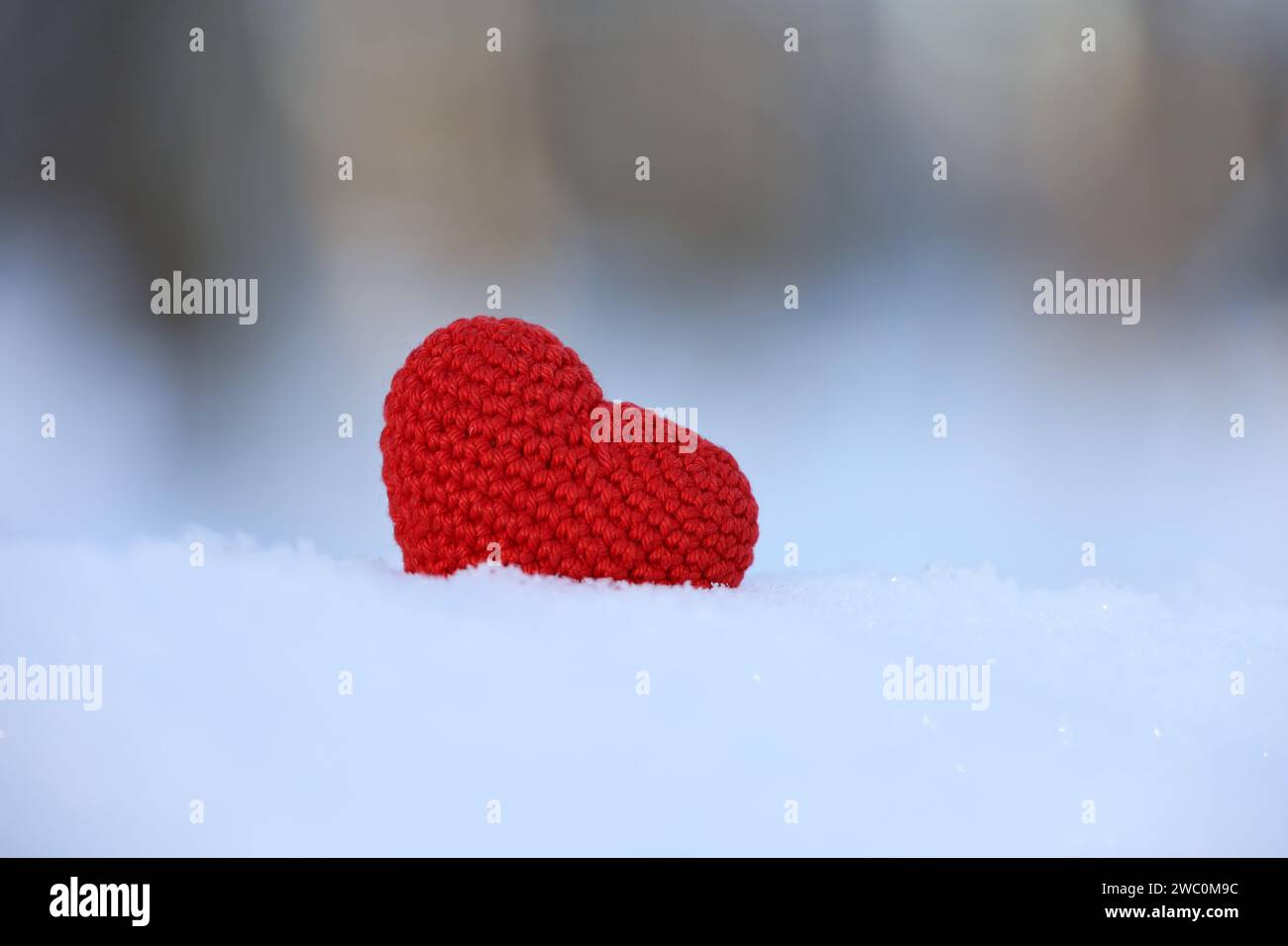 Rotes gestricktes Herz im Schnee auf verschwommenem Waldhintergrund. Symbol der romantischen Liebe, Valentinskarte Stockfoto
