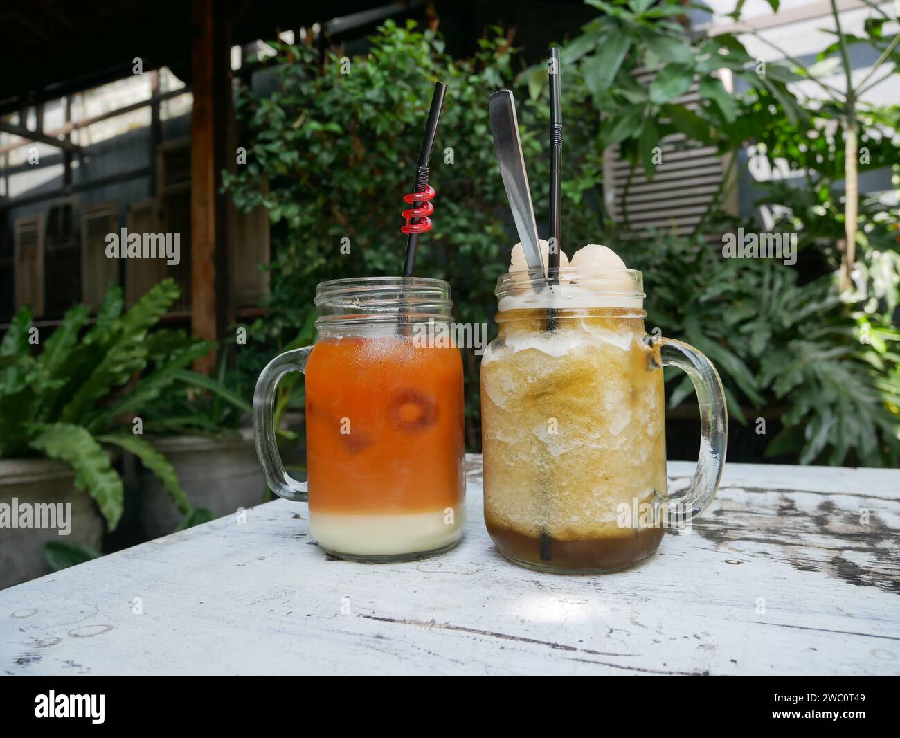 Eisgekühlter Litschi-Tee und eisgekühlter Thai-Tee serviert auf einem weißen Tisch mit tropischen Pflanzen und altem rustikalem Teakholz. Stockfoto