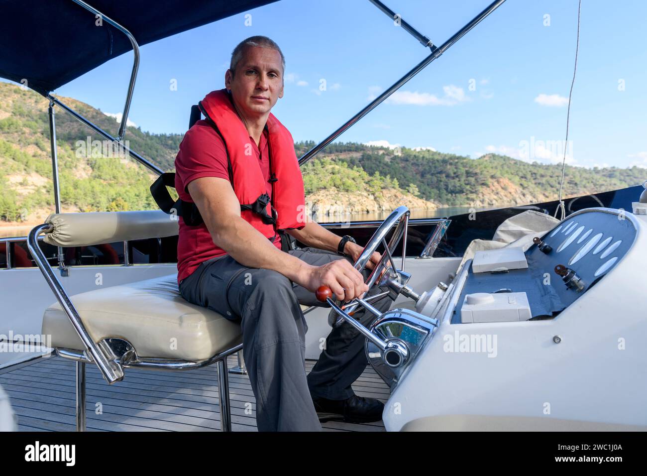 Junger kaukasischer Mann, der Motoryacht steuert. Kapitän des Motorbootes. Kapitän der Yacht. Segelkonzept. Mann mit aufblasbarer Schwimmweste. Stockfoto