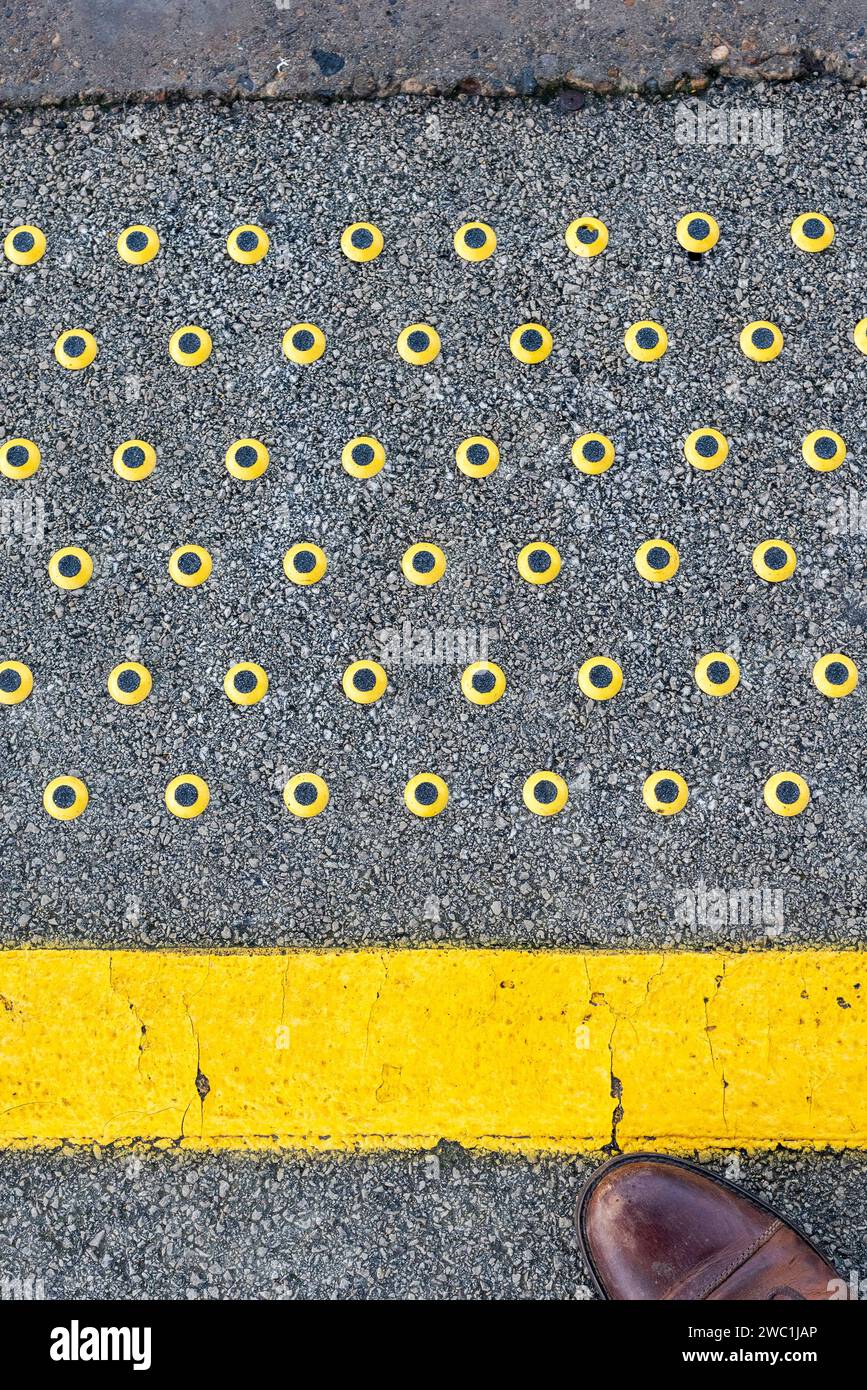 Taktiler Warnstreifen, gelbe Linie mit schwarzen und gelben Nieten am Bahnsteigrand, Vereinigtes Königreich, mit einem Teil des Pendlerschuhs sichtbar Stockfoto