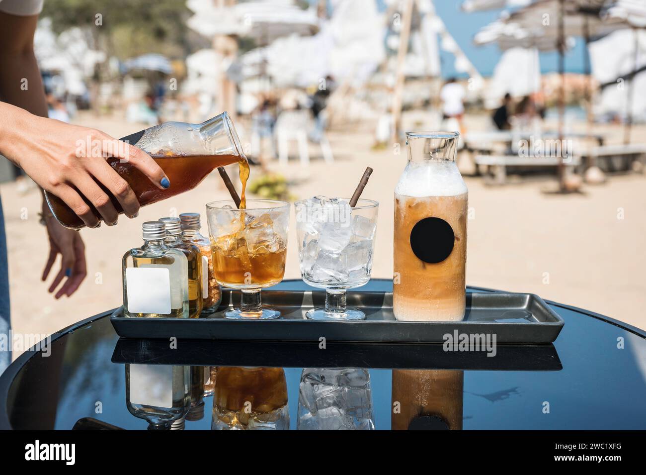 Schwarzer Kaffee von Hand aus der Flasche in Glas mit Eiswürfel und Eislatte, Sirup auf Tablett an sonnigem Tag Stockfoto