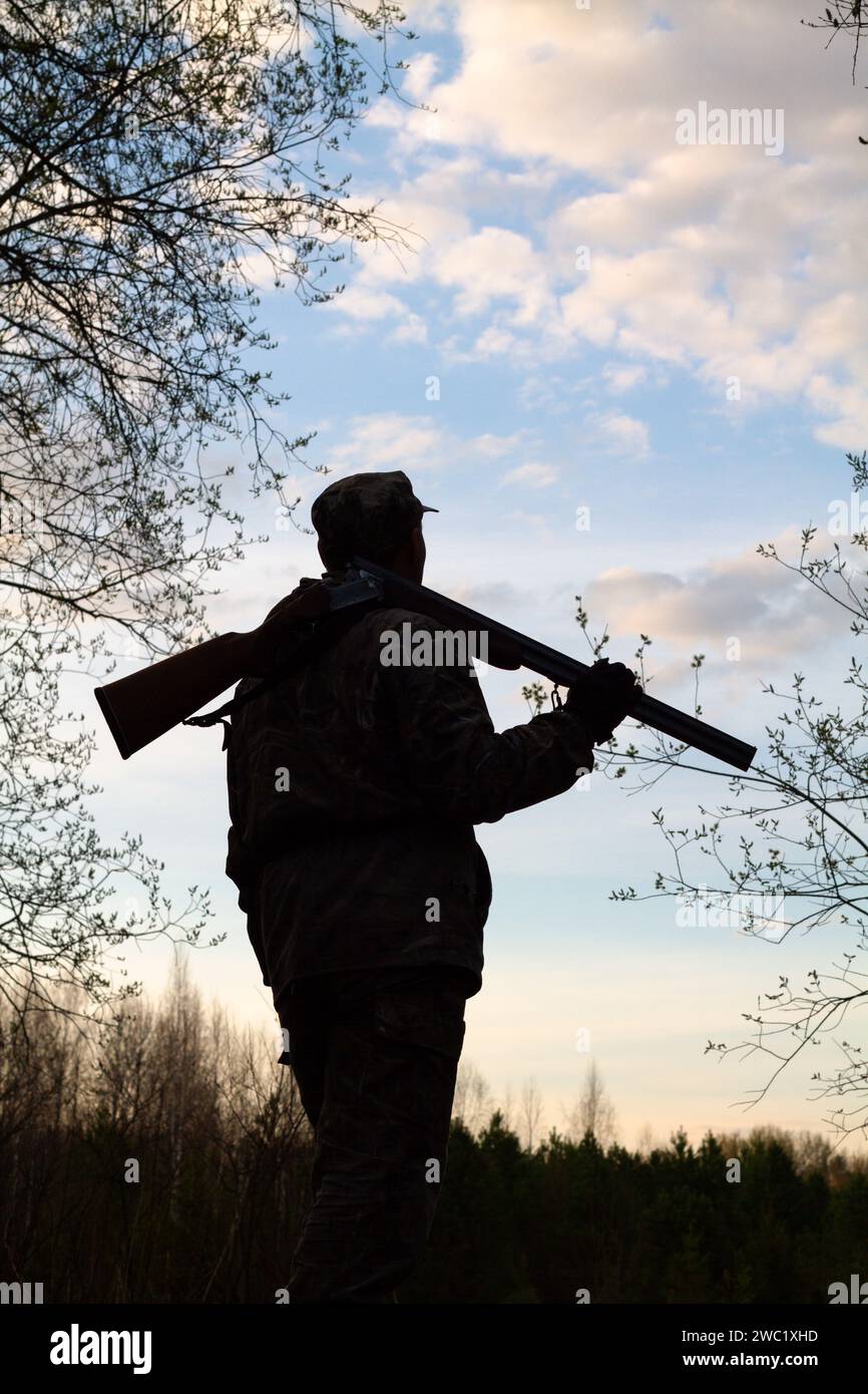 Eine schwarze Silhouette eines Jägers mit einer entladenen Schrotflinte auf der Schulter vor dem Hintergrund des Abendhimmels. Er steht und schaut sich den Dunkelheit an Stockfoto