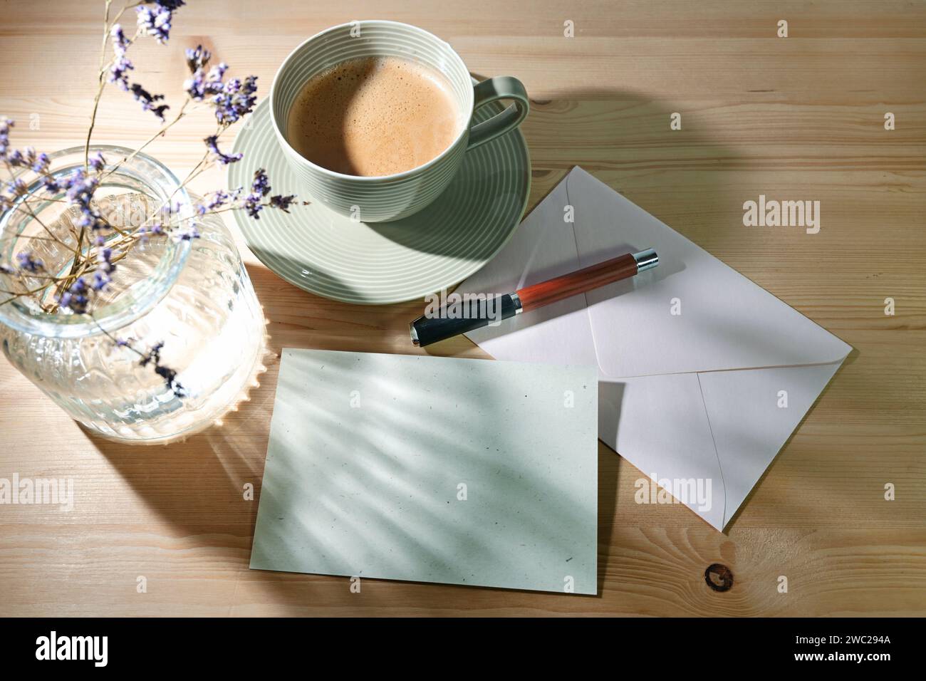Leere Grußkarte und Umschlag aus Naturpapier, Stift, Kaffeetasse und Glasvase mit sanften Sonnenlichtreflexionen auf einem Holztisch, pastellfarben Stockfoto
