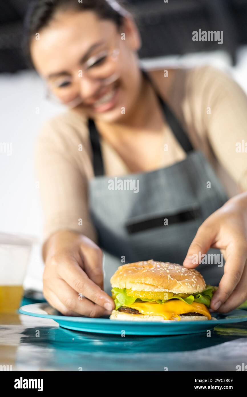 Junge Frau genießt und lächelt, während sie einen Hamburger zubereitet, Fast Food Restaurant, frische Zutaten. Geschäftsprodukt Stockfoto