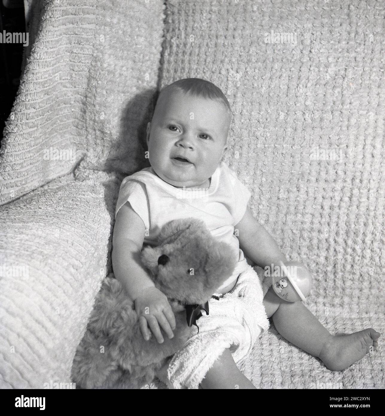 1950er Jahre, historisch, ein Kleinkind, das in der Ecke eines Baumwollsofas für ein Foto sitzt, mit Teddybär und Spielzeugrassel, England, Großbritannien. Stockfoto