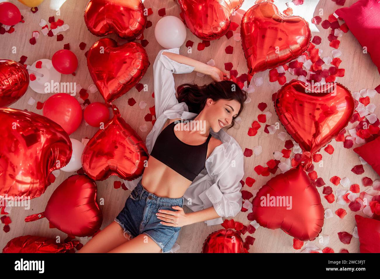 Blick von oben auf die junge Frau, die am Valentinstag auf Holzboden liegt und von roten Herzballons umgeben ist. Das Mädchen lächelt und trägt ein lässiges Outfit. Boden Stockfoto
