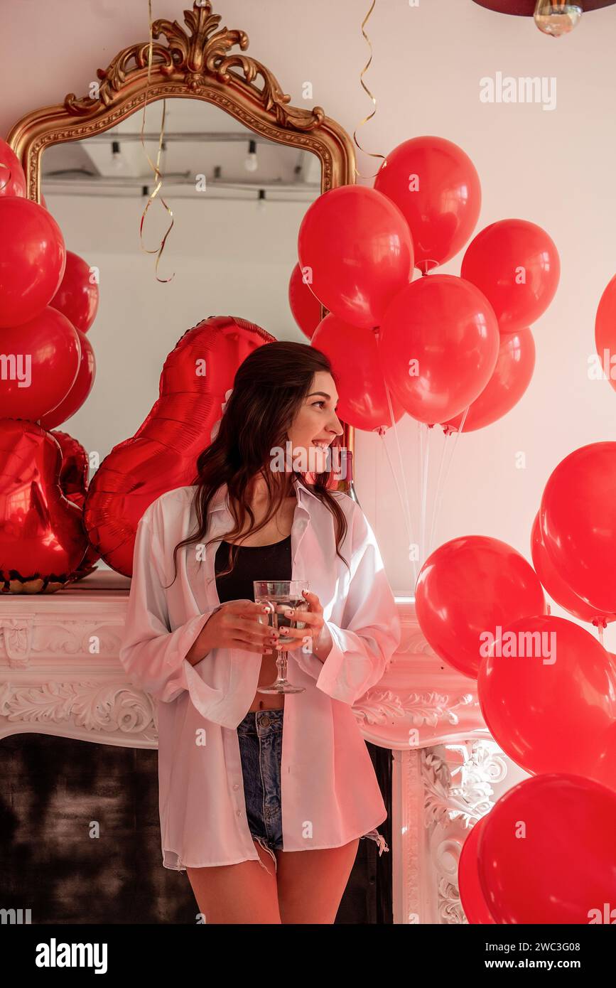 Junge Frau, die im Zimmer steht, gefüllt mit einer Mischung aus roten Ballons verschiedener Formen, mit einem romantischen weißen Kamin im Hintergrund. Mädchen, das Glas hält Stockfoto