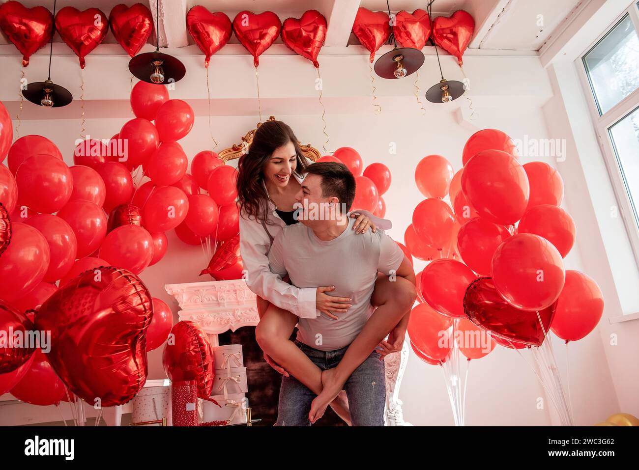 Verspielter, romantischer Moment zwischen verrücktem Paar in der Nähe von roten Ballons in einem hellen Raum mit weißer Inneneinrichtung. Frau sitzt auf dem Rücken des Mannes. Ein paar, das herumspielt Stockfoto