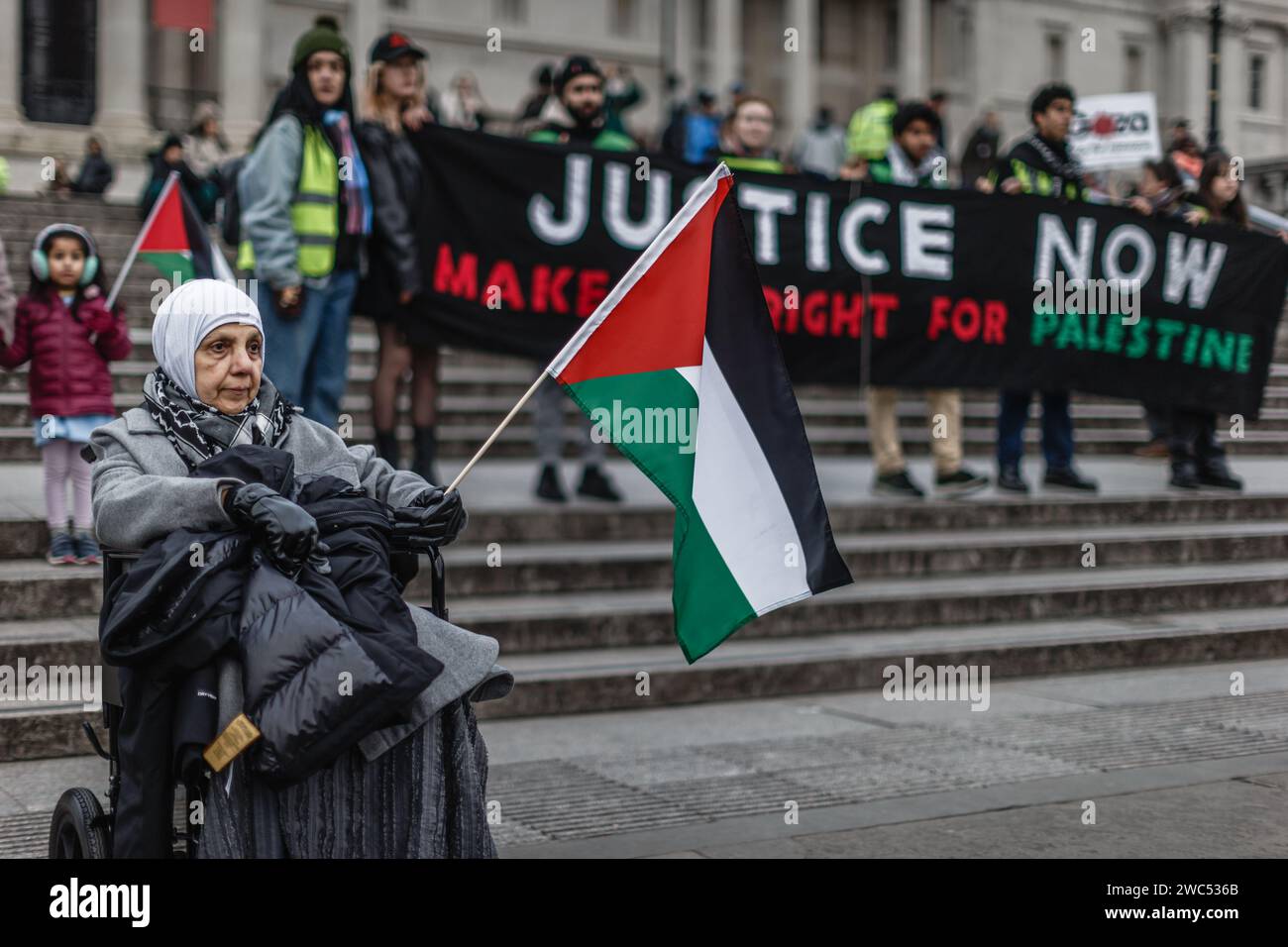 Eine alte Dame im Rollstuhl protestiert vor einem riesigen Banner "GERECHTIGKEIT macht es JETZT richtig für Palästina" auf dem Trafalgar Square. Stockfoto