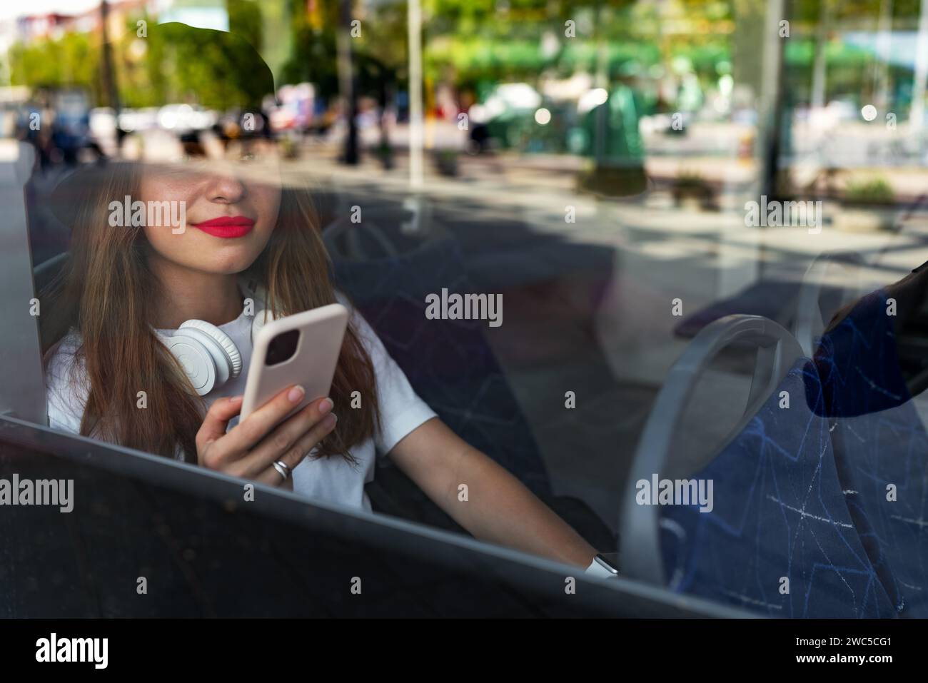 Urbane junge, stilvolle Frau mit roten Lippen, die ihr Smartphone benutzt, während sie im Bus unterwegs ist. Straßenfoto durch das Fenster. Stockfoto
