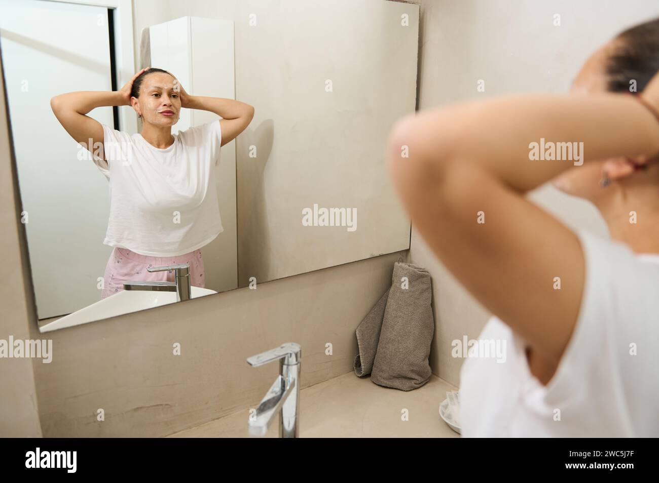 Reflexion im Badezimmerspiegel einer jungen, hübschen Frau mit strahlender, gesunder Haut, die sich lächelnd anschaut, während sie morgens einen Spa-Profi macht Stockfoto