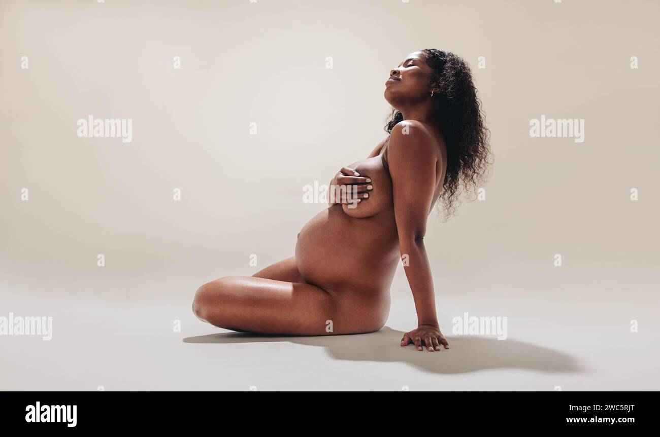 Selbstbewusste Afrikanerin sitzt in einem Studio und zeigt ihren dunkelhäutigen, nackten schwangeren Körper. Junge schwarze Frau, die ihre Körperpositivität und natura annimmt Stockfoto