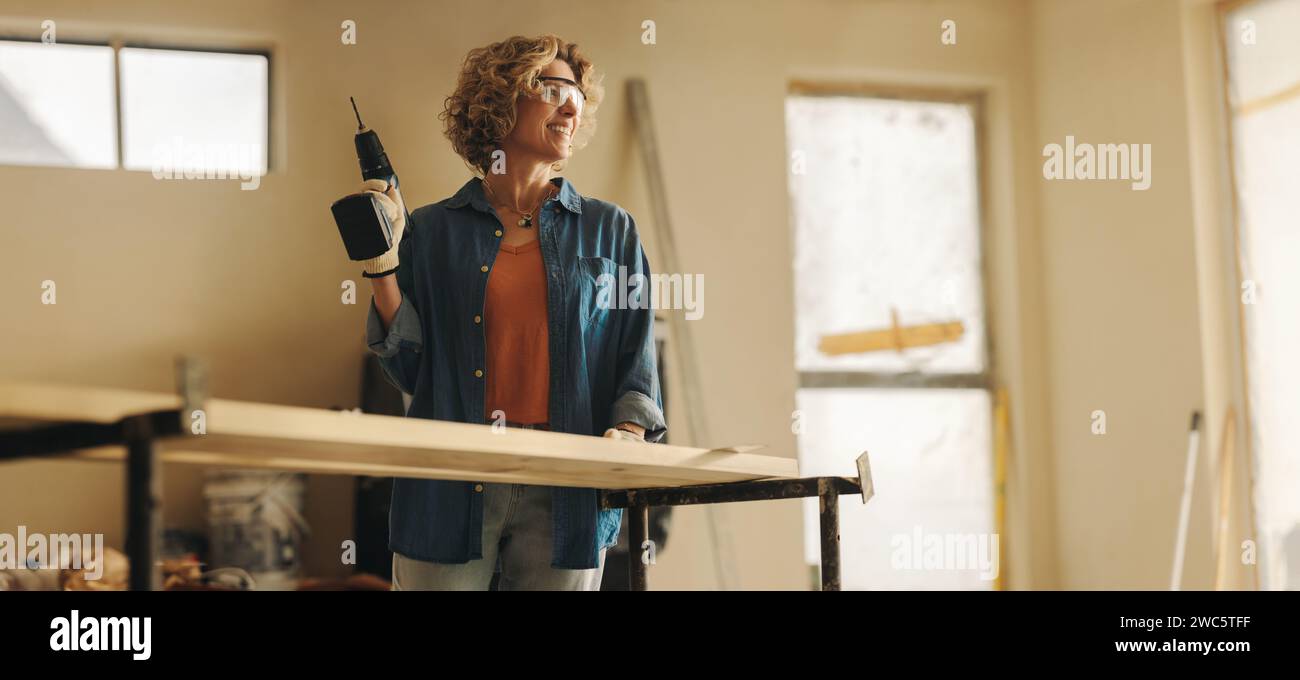 Glückliche weiße Frau renoviert die Küche ihres Hauses. Mit Handwerkzeugen und Schutzbrille montiert sie die Sockelleisten geschickt mit einer Bohrpistole. Stockfoto