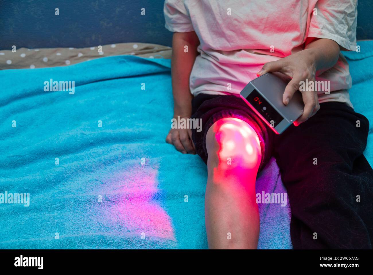 Nahaufnahme der Behandlung von Beinschmerzen mit dem Rotlichttherapie-Handgerät zu Hause auf dem Bett Stockfoto