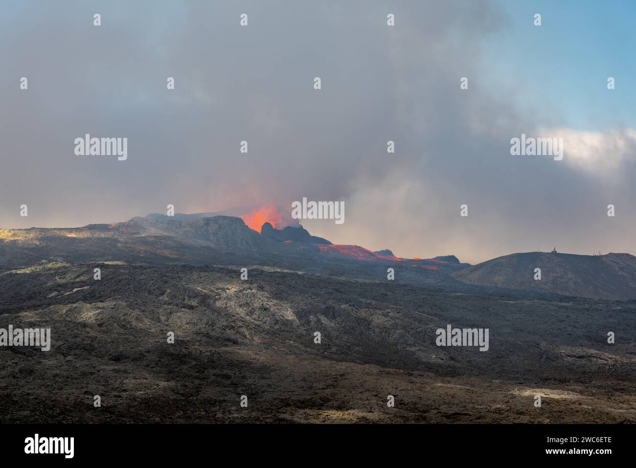 Blick über ein gehärtetes schwarzgraues Lavafeld. Durch schimmernde heiße Luft brechen Lava und Rauch aus einem Vulkan in der Ferne aus. Stockfoto