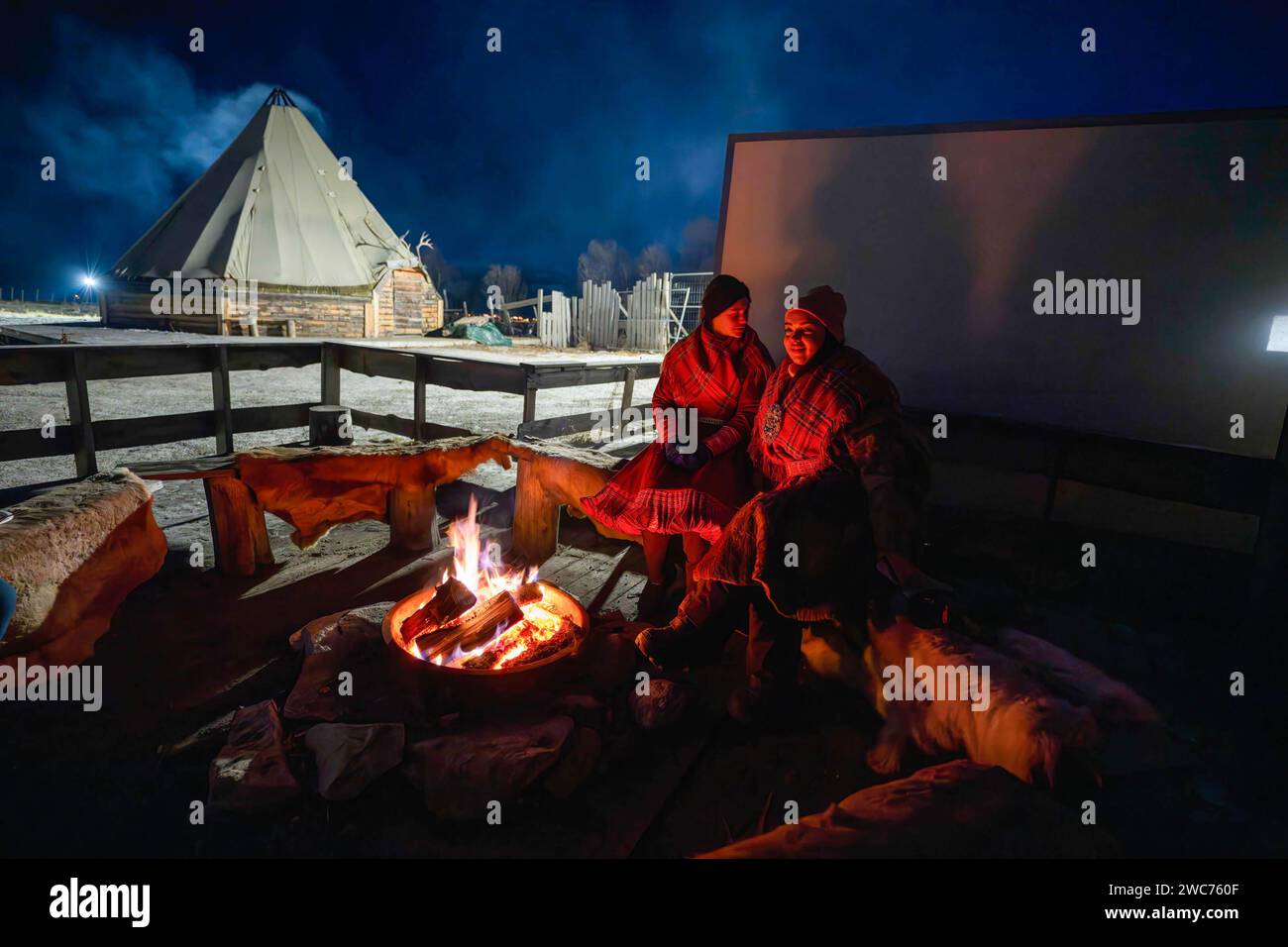 Zwei samische Frauen teilen sich ein Feuer an einem Lagerfeuer vor einem Gemeinschaftshaus auf einer samischen Farm. In Tromsø und Umgebung ist die Rentierhaltung eine wichtige Tätigkeit. Es gibt mehrere Rentierfarmen, die Besuchern Erfahrungen bieten und die Gelegenheit bieten, mehr über das Rentierleben und die Kultur der Sami zu erfahren. Die Rentierhaltung in Norwegen ist eine nachhaltige Tätigkeit, die unter Beachtung traditioneller Praktiken durchgeführt wird und zur Erhaltung der samischen Kultur beiträgt. Außerdem spielen Rentiere eine wichtige Rolle im arktischen Ökosystem und sind ein ikonisches Symbol der Region. Stockfoto