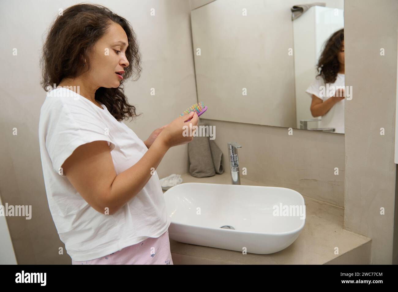 Verärgerte Frau, die verlorene Haare von einer Haarbürste entfernt, nachdem sie ihr geschädigtes lockiges und trockenes Haar gekämmt hat und im Badezimmer zu Hause am Waschbecken steht. Haarpflegekonzept Stockfoto
